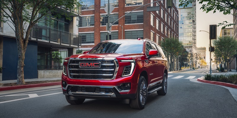 A shining red 2026 GMC Yukon SUV is driving down the street toward us in the business district of the city.